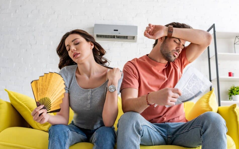 Frustrated couple fanning themselves in hot room with broken air conditioner